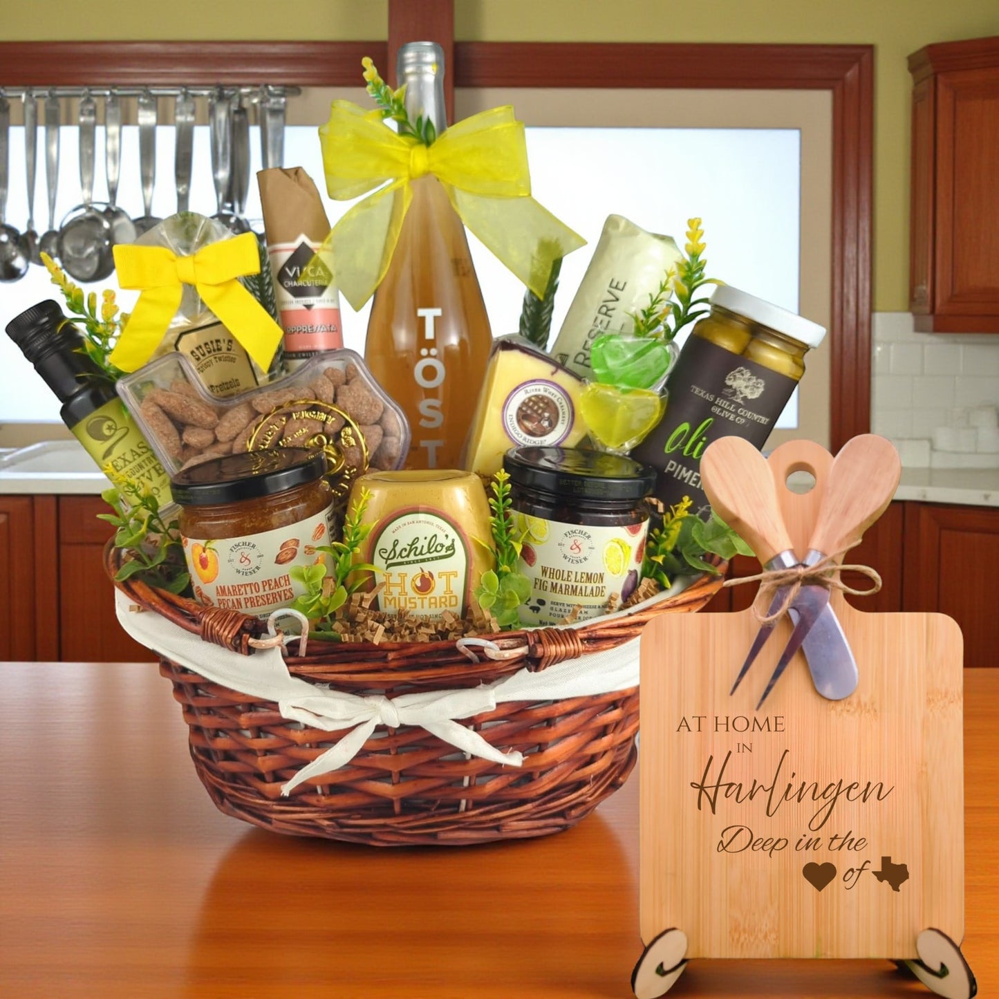 Gift basket filled with Texas treats, preserves, mustard, cheese, sparkling drink, and snacks, with a bamboo board engraved “At Home in Harlingen, Deep in the Heart of Texas” displayed in a kitchen.