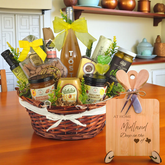 Gift basket filled with Texas treats, preserves, mustard, cheese, sparkling drink, and snacks, with a bamboo board engraved “At Home in Midland, Deep in the Heart of Texas” displayed in a kitchen.