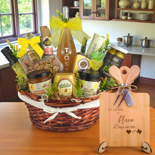 Gift basket filled with Texas treats, preserves, mustard, cheese, sparkling drink, and snacks, with a bamboo board engraved “At Home in Plano, Deep in the Heart of Texas” displayed in a kitchen.