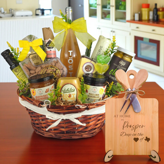Gift basket filled with Texas treats, preserves, mustard, cheese, sparkling drink, and snacks, with a bamboo board engraved “At Home in Prosper, Deep in the Heart of Texas” displayed in a kitchen.