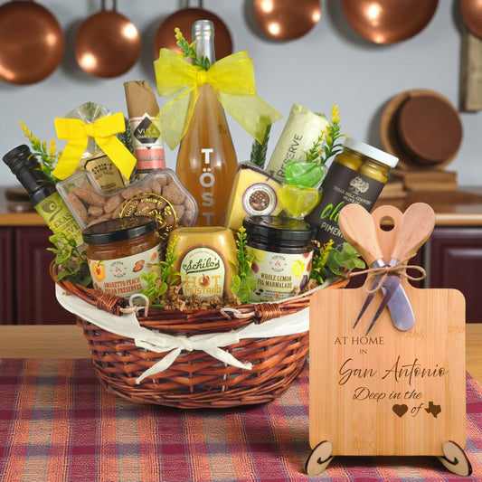 Gift basket filled with Texas treats, preserves, mustard, cheese, sparkling drink, and snacks, with a bamboo board engraved “At Home in San Antonio, Deep in the Heart of Texas” displayed in a kitchen.