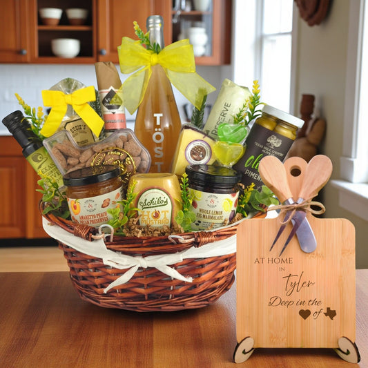 Gift basket filled with Texas treats, preserves, mustard, cheese, sparkling drink, and snacks, with a bamboo board engraved “At Home in Tyler, Deep in the Heart of Texas” displayed in a kitchen.