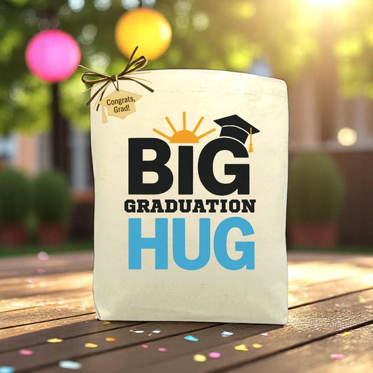 A canvas gift tote with 'BIG GRADUATION HUG' printed in large blue and black letters, featuring a graduation cap icon and a sun rise graphic. The tote is tied with a ribbon and placed on a table with blurred colorful balloons and decorations in the background.