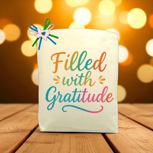 Canvas tote bag printed with the phrase “Filled with Gratitude” in colorful script, accented with a ribbon bow and Texas charm, displayed on a wooden table with warm light bokeh background.