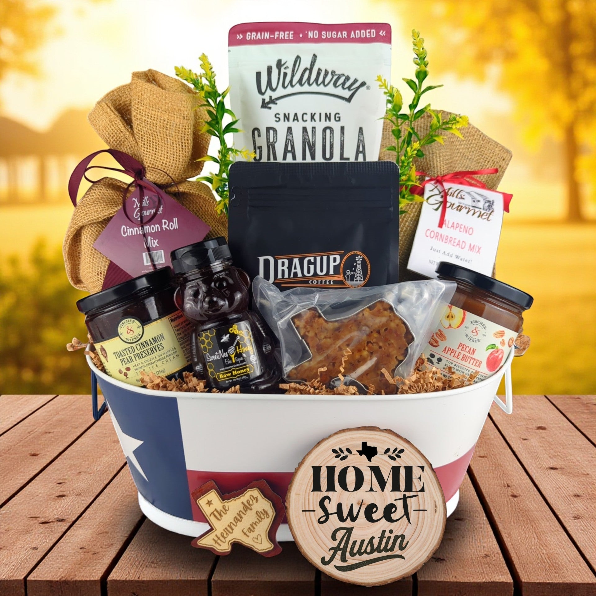 Austin Home Breakfast Basket with rustic outdoor background, Texas tin filled with pecan coffee, cinnamon roll mix, jalapeño cornbread mix, granola, honey, praline, and preserves.