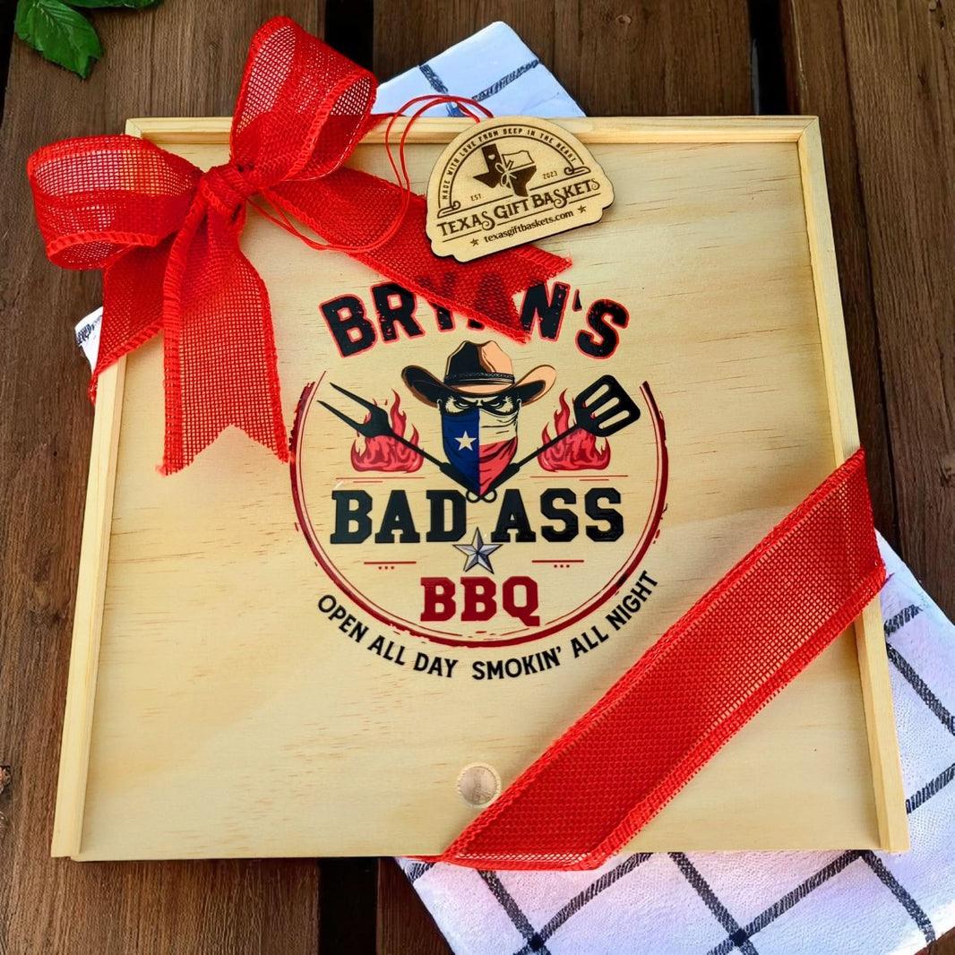 Wooden gift box with 'Texas Gift Baskets' wooden tag and 'Bad Ass BBQ' design and red ribbon on a wooden surface.
