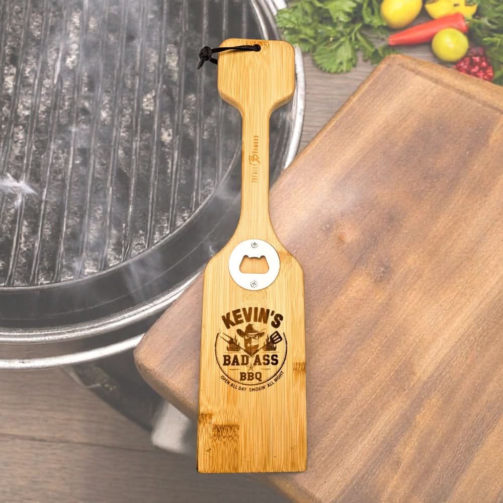 a bamboo bbq scraper with "Kevin's bad ass bbq" engraved on it, on an outdoor cutting board next to a grill