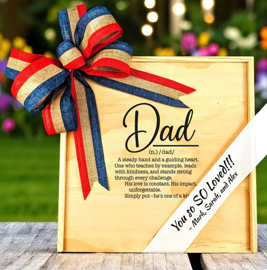 Wooden gift box with 'definition of dad' wording and a personalized ribbon on top, accented by a red, white, and blue bow.
