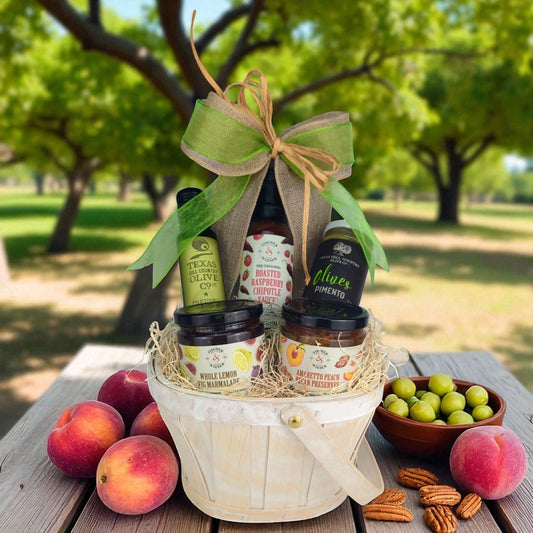 a texas hill country themed gift basket with fischer & weiser products and texas hill country olive co. products, on a wooden table next to peaches, pecans and olives