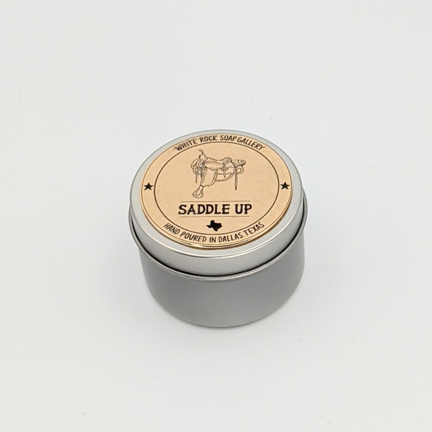Silver metal candle tin labeled “Saddle Up” with a western saddle illustration, hand-poured in Dallas, Texas, photographed on a clean white background.