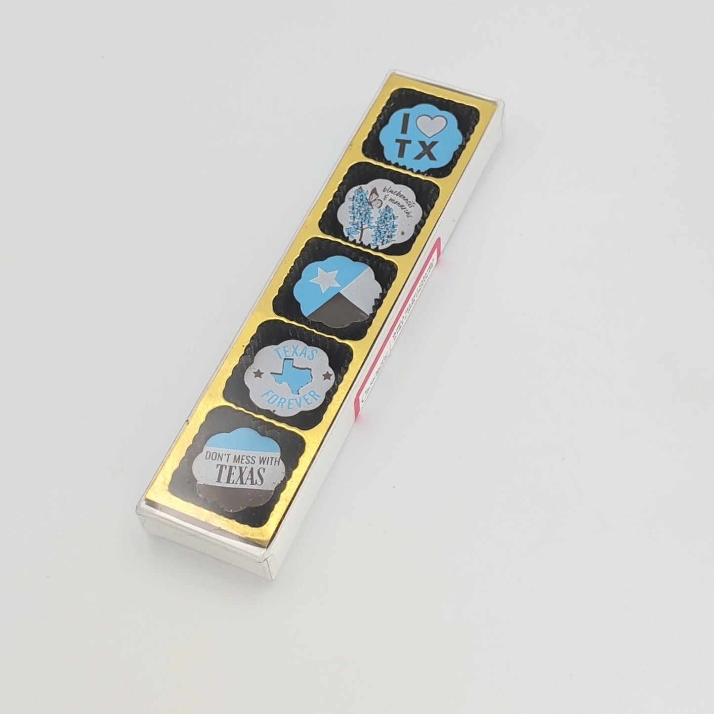 Slim rectangular clear gift box displaying five Texas-themed chocolate-covered caramels with designs including “I Love TX,” bluebonnets, the Texas flag, “Texas Forever,” and “Don’t Mess With Texas,” arranged on a gold backing against a white background.
