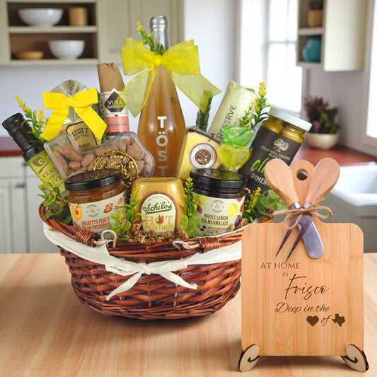 Gift basket filled with Texas treats, preserves, mustard, cheese, sparkling drink, and snacks, with a bamboo board engraved “At Home in Frisco, Deep in the Heart of Texas” displayed in a kitchen.