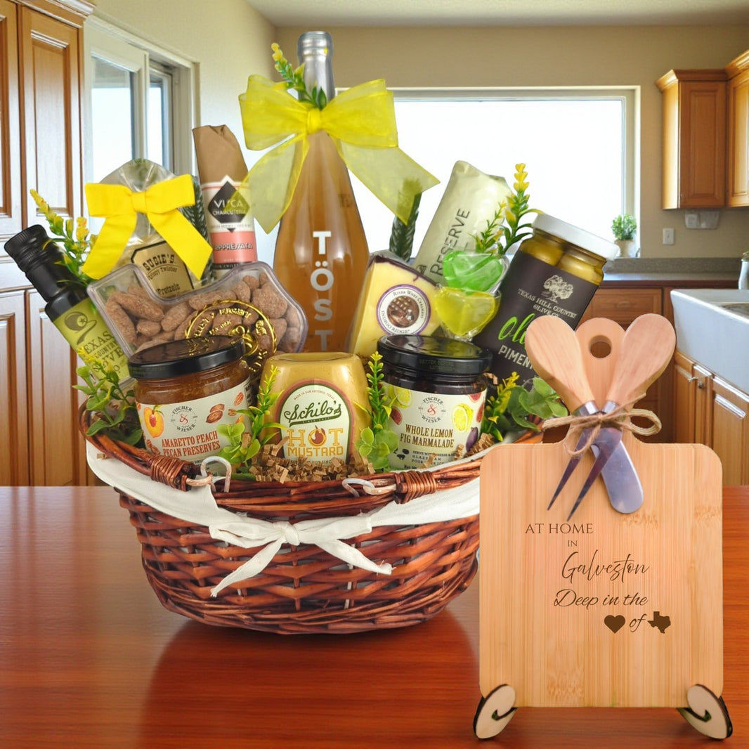 Gift basket filled with Texas treats, preserves, mustard, cheese, sparkling drink, and snacks, with a bamboo board engraved “At Home in Galveston, Deep in the Heart of Texas” displayed in a kitchen.