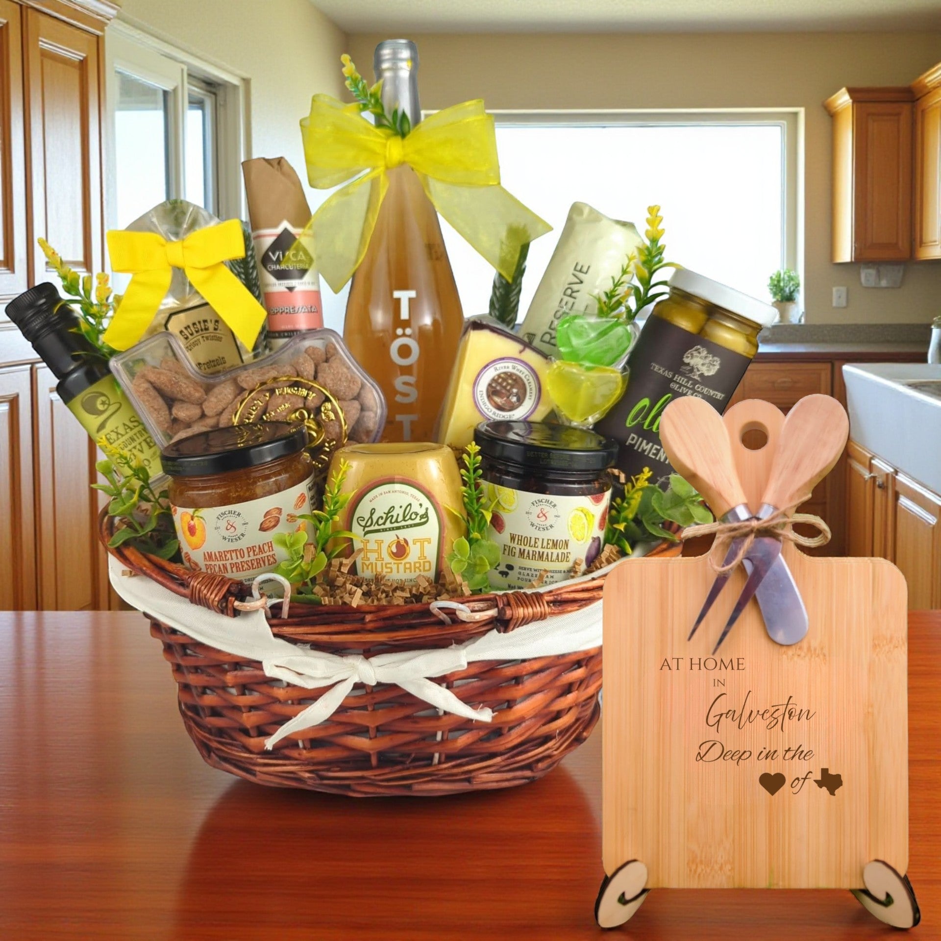 Gift basket filled with Texas treats, preserves, mustard, cheese, sparkling drink, and snacks, with a bamboo board engraved “At Home in Galveston, Deep in the Heart of Texas” displayed in a kitchen.