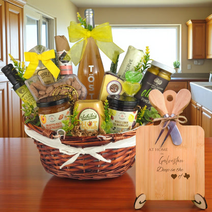 Gift basket filled with Texas treats, preserves, mustard, cheese, sparkling drink, and snacks, with a bamboo board engraved “At Home in Galveston, Deep in the Heart of Texas” displayed in a kitchen.