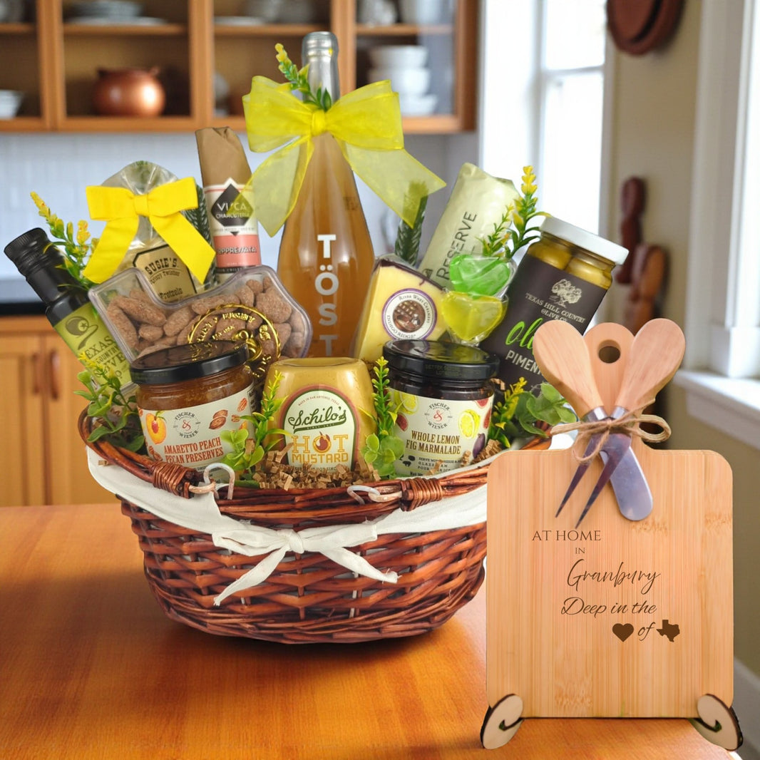 Gift basket filled with Texas treats, preserves, mustard, cheese, sparkling drink, and snacks, with a bamboo board engraved “At Home in Granbury, Deep in the Heart of Texas” displayed in a kitchen.