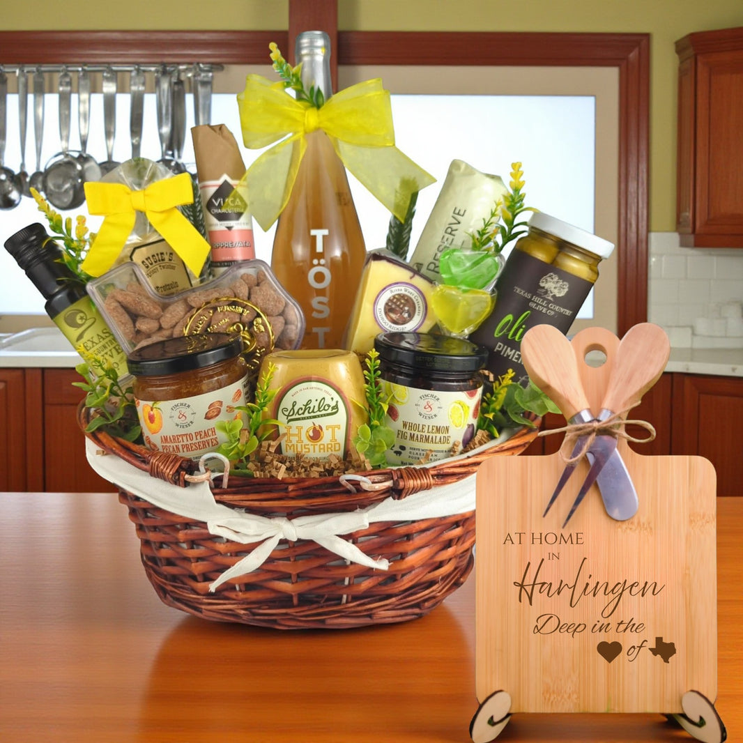 Gift basket filled with Texas treats, preserves, mustard, cheese, sparkling drink, and snacks, with a bamboo board engraved “At Home in Harlingen, Deep in the Heart of Texas” displayed in a kitchen.