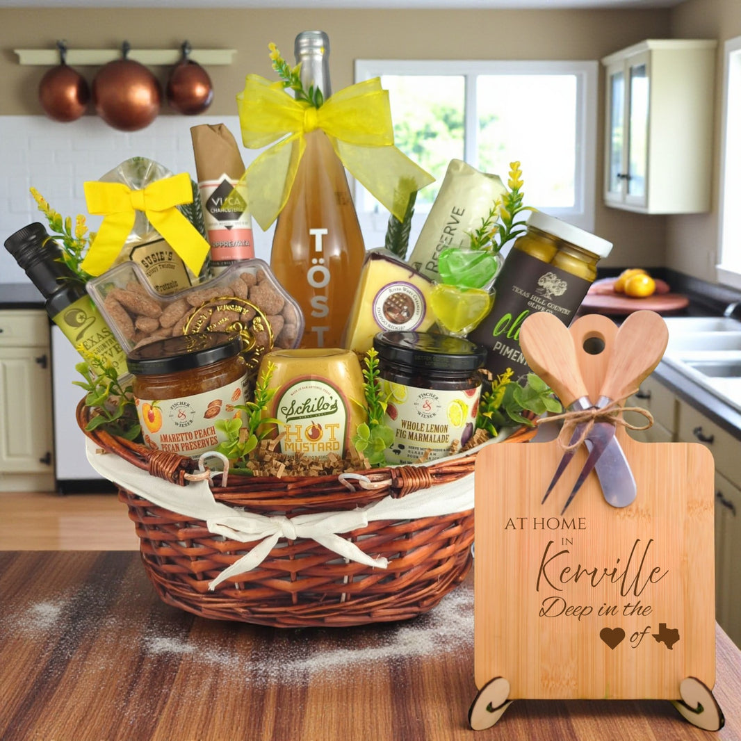 Gift basket filled with Texas treats, preserves, mustard, cheese, sparkling drink, and snacks, with a bamboo board engraved “At Home in Kerrville, Deep in the Heart of Texas” displayed in a kitchen.