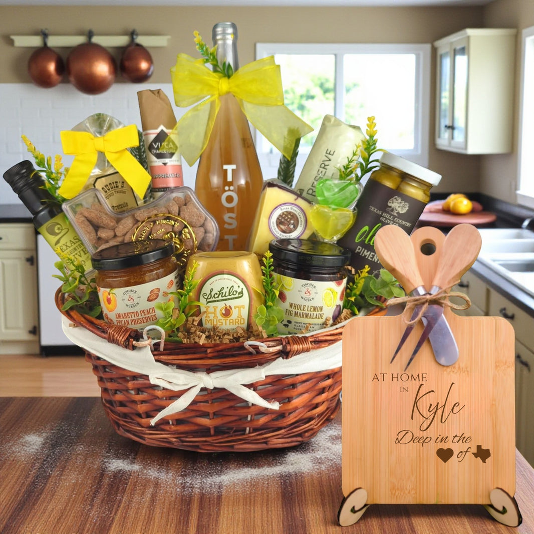 Gift basket filled with Texas treats, preserves, mustard, cheese, sparkling drink, and snacks, with a bamboo board engraved “At Home in Kyle, Deep in the Heart of Texas” displayed in a kitchen.