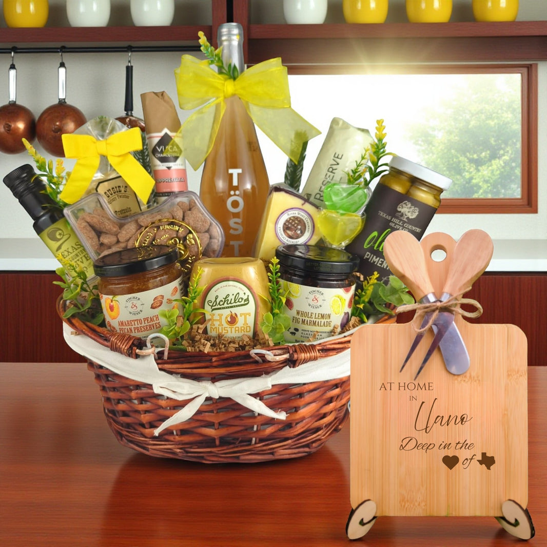 Gift basket filled with Texas treats, preserves, mustard, cheese, sparkling drink, and snacks, with a bamboo board engraved “At Home in Llano, Deep in the Heart of Texas” displayed in a kitchen.