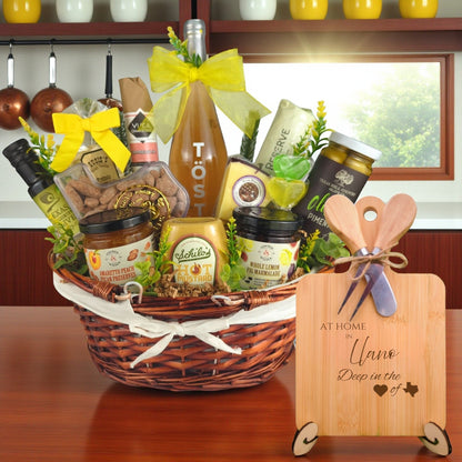 Gift basket filled with Texas treats, preserves, mustard, cheese, sparkling drink, and snacks, with a bamboo board engraved “At Home in Llano, Deep in the Heart of Texas” displayed in a kitchen.