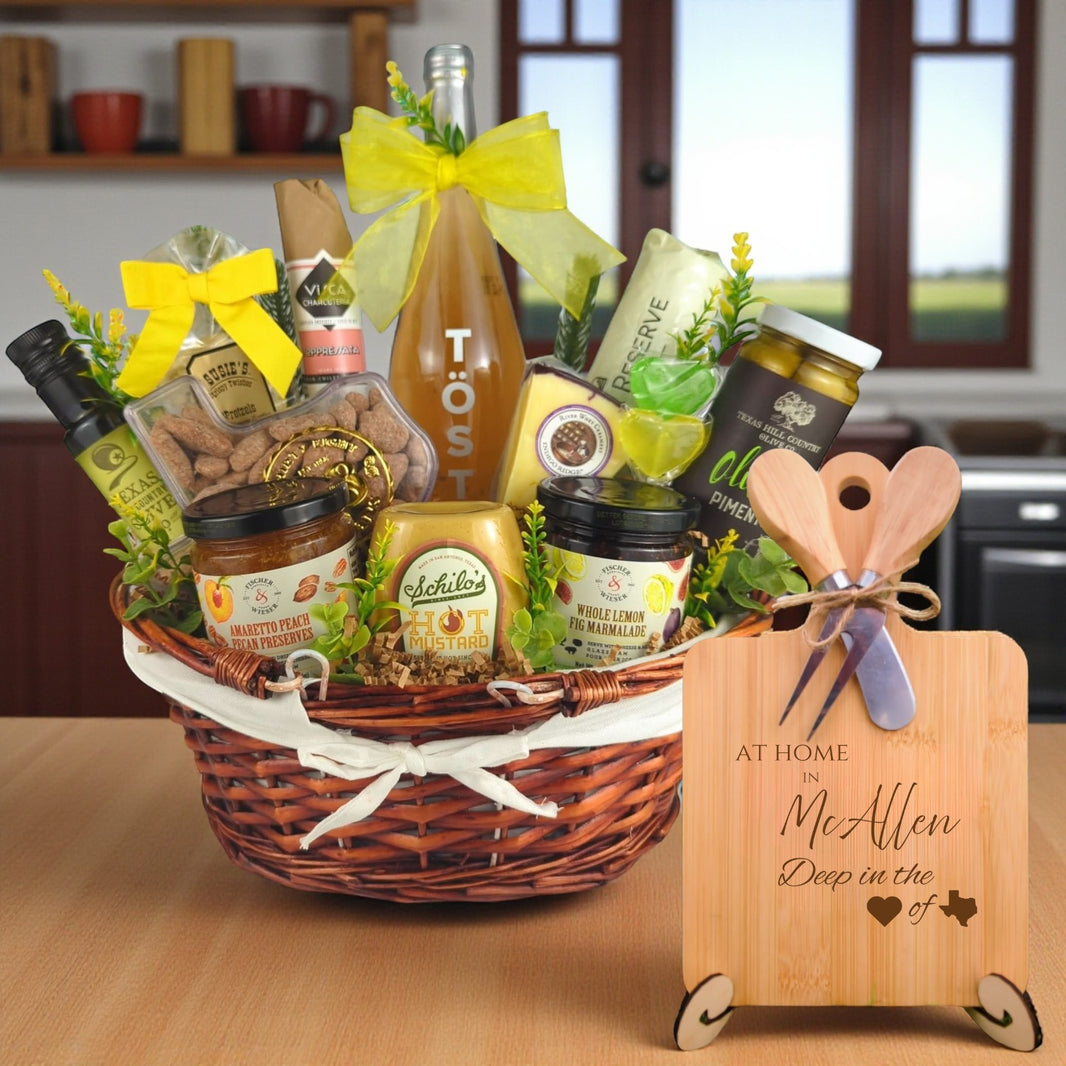 Gift basket filled with Texas treats, preserves, mustard, cheese, sparkling drink, and snacks, with a bamboo board engraved “At Home in McAllen, Deep in the Heart of Texas” displayed in a kitchen.