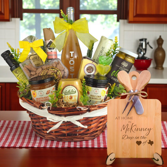 Gift basket filled with Texas treats, preserves, mustard, cheese, sparkling drink, and snacks, with a bamboo board engraved “At Home in McKinney, Deep in the Heart of Texas” displayed in a kitchen.