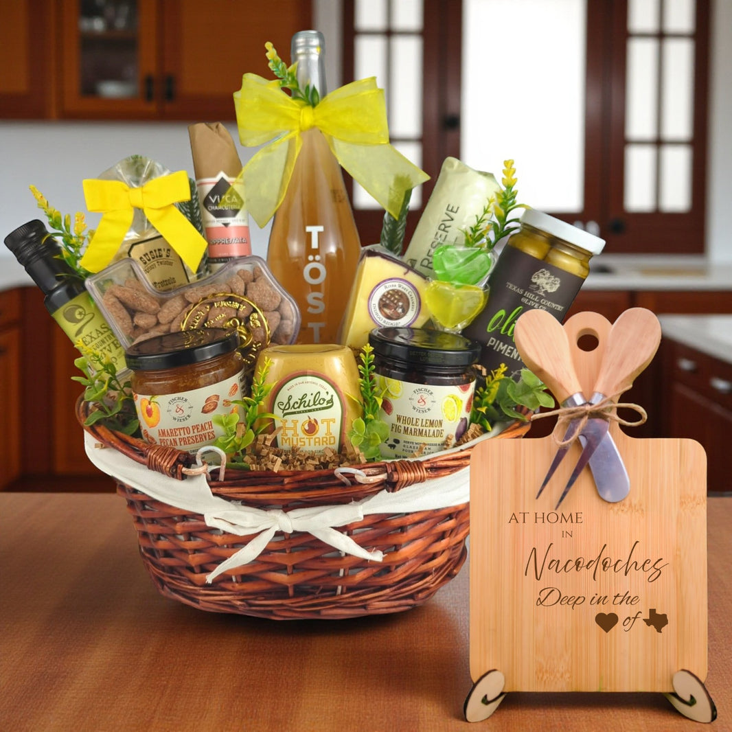 Gift basket filled with Texas treats, preserves, mustard, cheese, sparkling drink, and snacks, with a bamboo board engraved “At Home in Nacogdoches, Deep in the Heart of Texas” displayed in a kitchen.