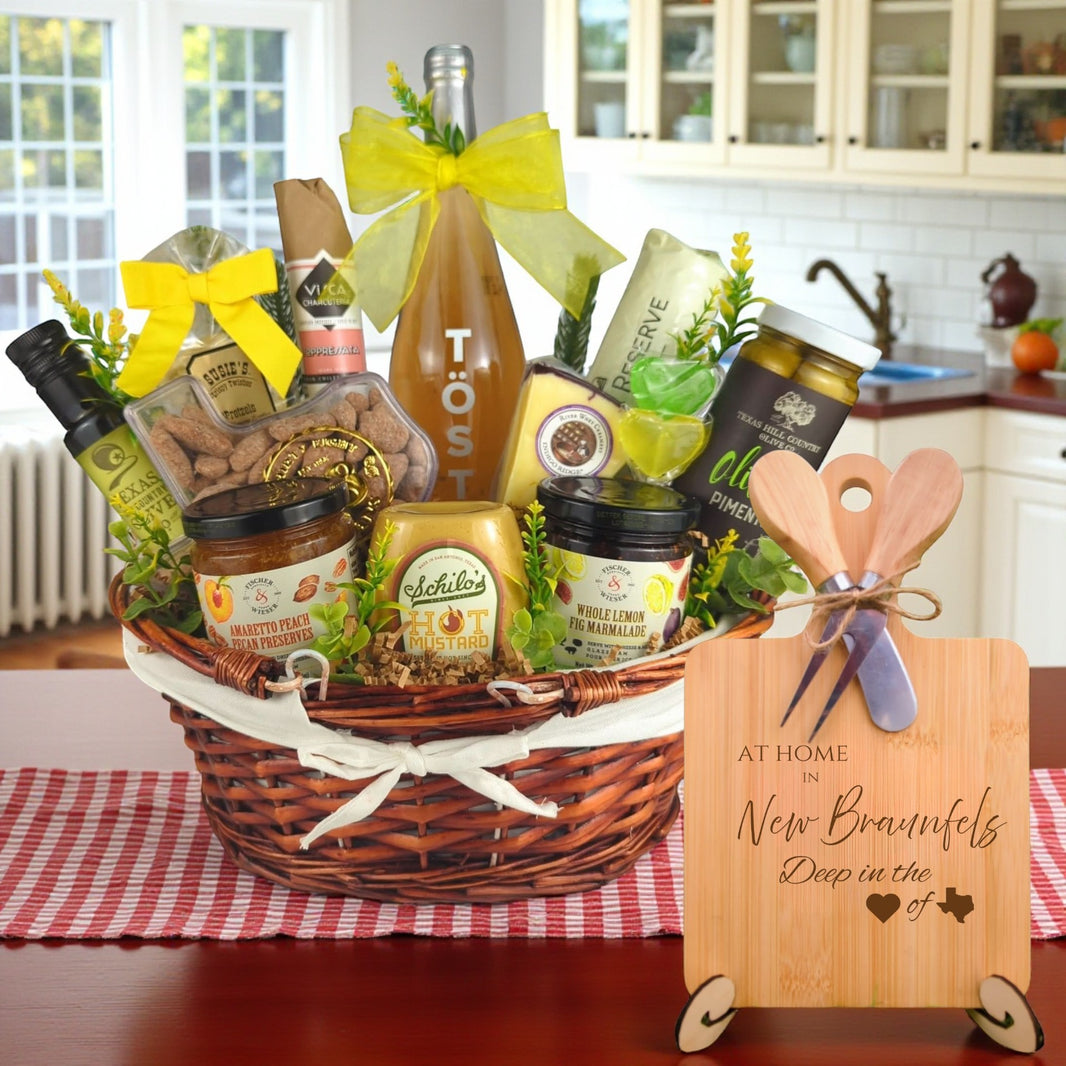 Gift basket filled with Texas treats, preserves, mustard, cheese, sparkling drink, and snacks, with a bamboo board engraved “At Home in New Braunfels, Deep in the Heart of Texas” displayed in a kitchen.