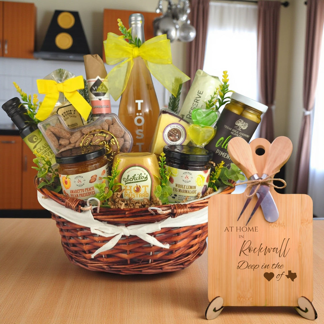 Gift basket filled with Texas treats, preserves, mustard, cheese, sparkling drink, and snacks, with a bamboo board engraved “At Home in Rockwall, Deep in the Heart of Texas” displayed in a kitchen.