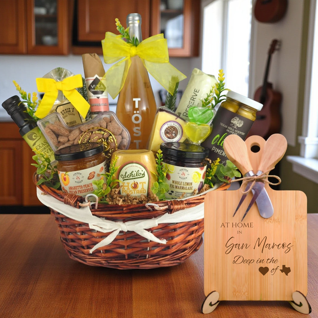 Gift basket filled with Texas treats, preserves, mustard, cheese, sparkling drink, and snacks, with a bamboo board engraved “At Home in San Marcos, Deep in the Heart of Texas” displayed in a kitchen.