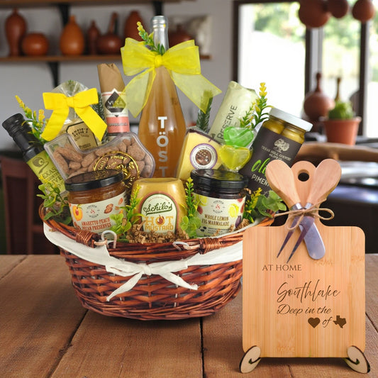 Gift basket filled with Texas treats, preserves, mustard, cheese, sparkling drink, and snacks, with a bamboo board engraved “At Home in Southlake, Deep in the Heart of Texas” displayed in a kitchen.