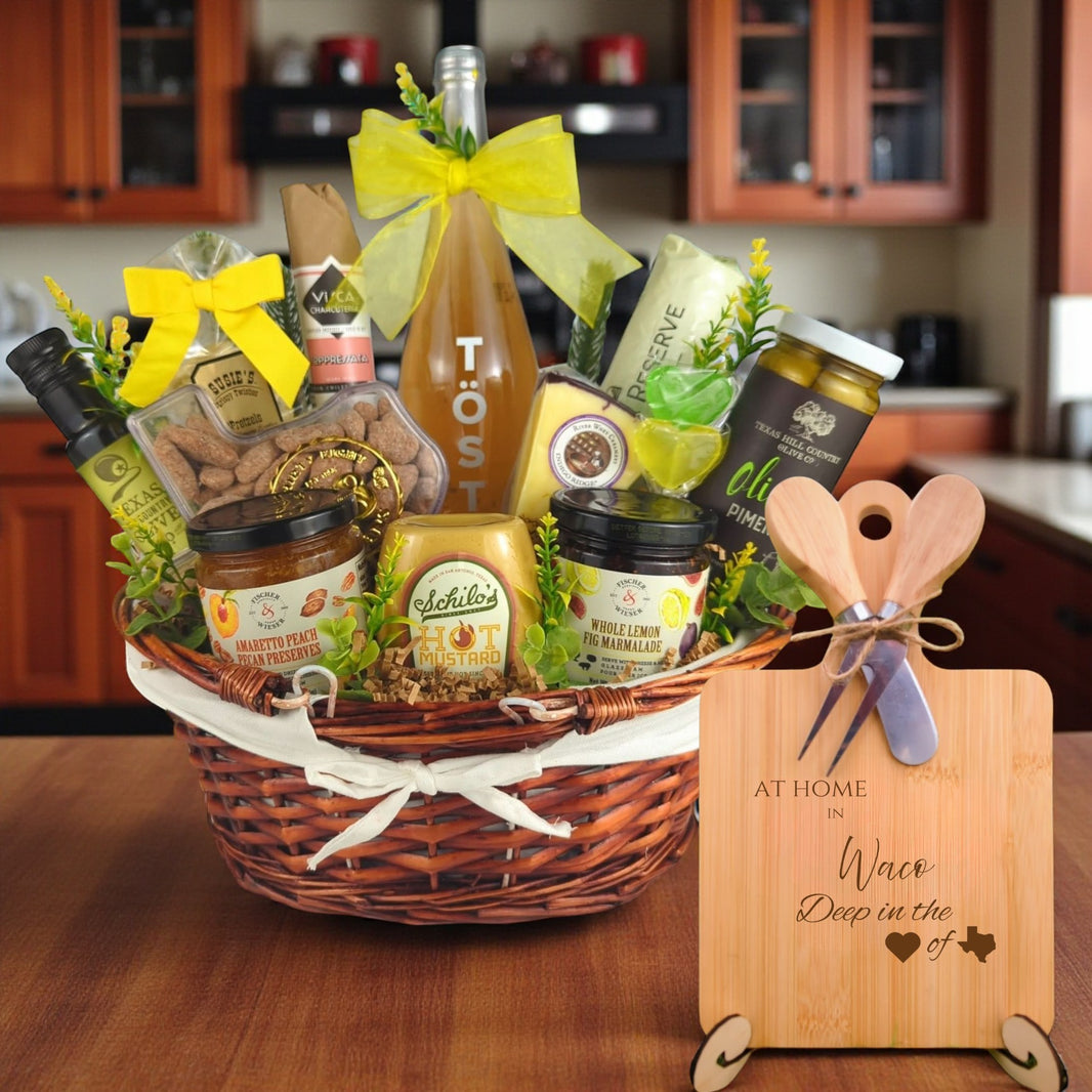 Gift basket filled with Texas treats, preserves, mustard, cheese, sparkling drink, and snacks, with a bamboo board engraved “At Home in Waco, Deep in the Heart of Texas” displayed in a kitchen.