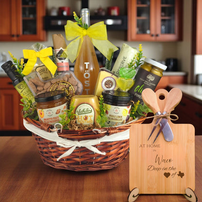 Gift basket filled with Texas treats, preserves, mustard, cheese, sparkling drink, and snacks, with a bamboo board engraved “At Home in Waco, Deep in the Heart of Texas” displayed in a kitchen.