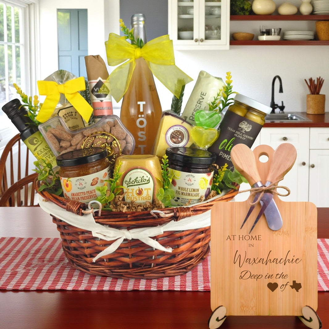 Gift basket filled with Texas treats, preserves, mustard, cheese, sparkling drink, and snacks, with a bamboo board engraved “At Home in Waxahachie, Deep in the Heart of Texas” displayed in a kitchen.