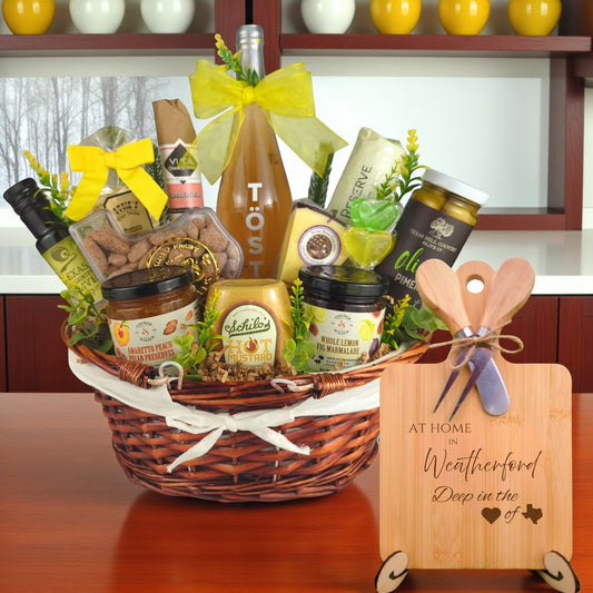Gift basket filled with Texas treats, preserves, mustard, cheese, sparkling drink, and snacks, with a bamboo board engraved “At Home in Weatherford, Deep in the Heart of Texas” displayed in a kitchen.