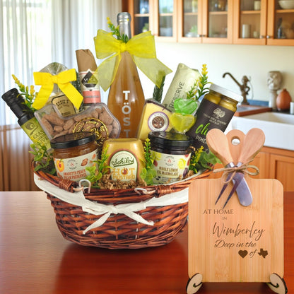 Gift basket filled with Texas treats, preserves, mustard, cheese, sparkling drink, and snacks, with a bamboo board engraved “At Home in Wimberley, Deep in the Heart of Texas” displayed in a kitchen.