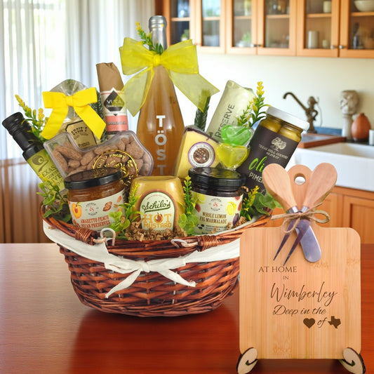 Gift basket filled with Texas treats, preserves, mustard, cheese, sparkling drink, and snacks, with a bamboo board engraved “At Home in Wimberley, Deep in the Heart of Texas” displayed in a kitchen.