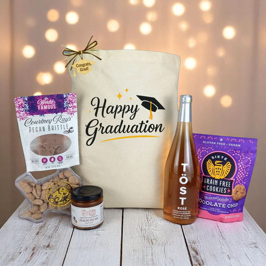 A gift tote with 'Happy Graduation' printed on it, accompanied by a variety of Texas-made treats including cookies, peacan butter, and non alcoholic rose, all displayed on a wooden surface with graduation caps and lights in the background.