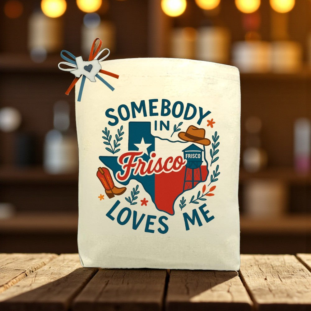 “Somebody In Frisco Loves Me” tote bag with Texas design, featuring cowboy boots, a cowboy hat, and Frisco landmarks, displayed on a wooden table with soft background lights.