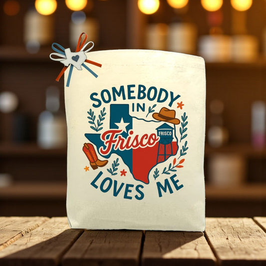 “Somebody In Frisco Loves Me” tote bag with Texas design, featuring cowboy boots, a cowboy hat, and Frisco landmarks, displayed on a wooden table with soft background lights.