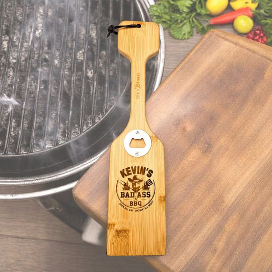 a bamboo bbq scraper with "Kevin's bad ass bbq" engraved on it, on an outdoor cutting board next to a grill 