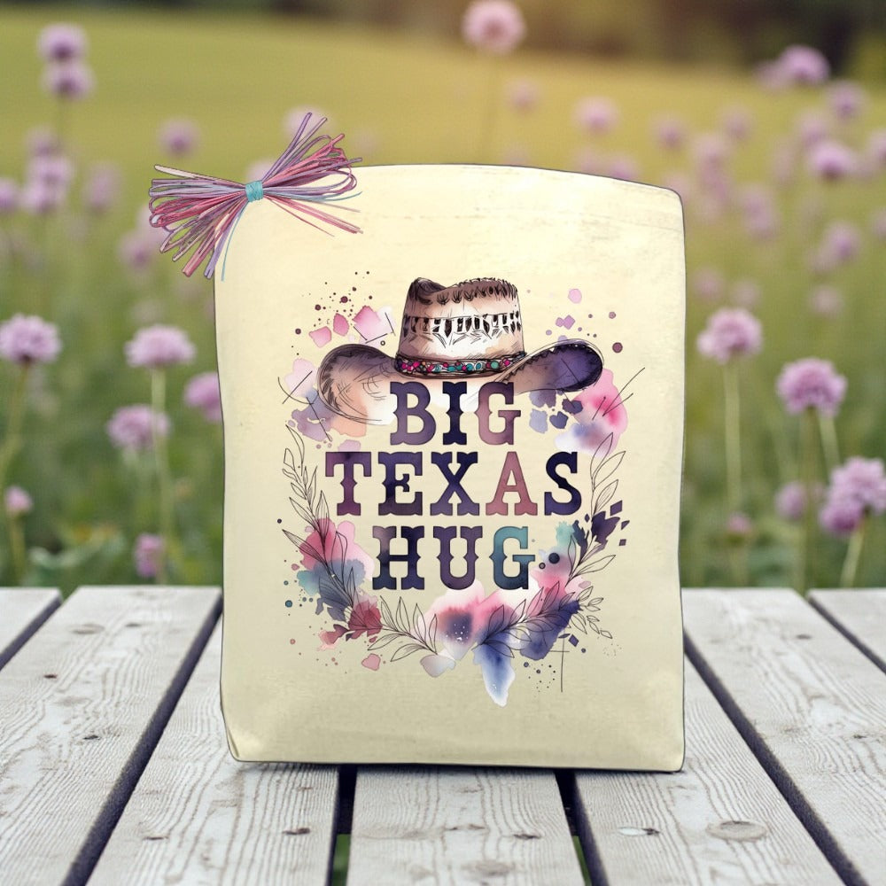 Big Texas Hug Cowgirl Edition gift tote with floral cowgirl hat design shown outdoors on a table in a Texas wildflower setting.