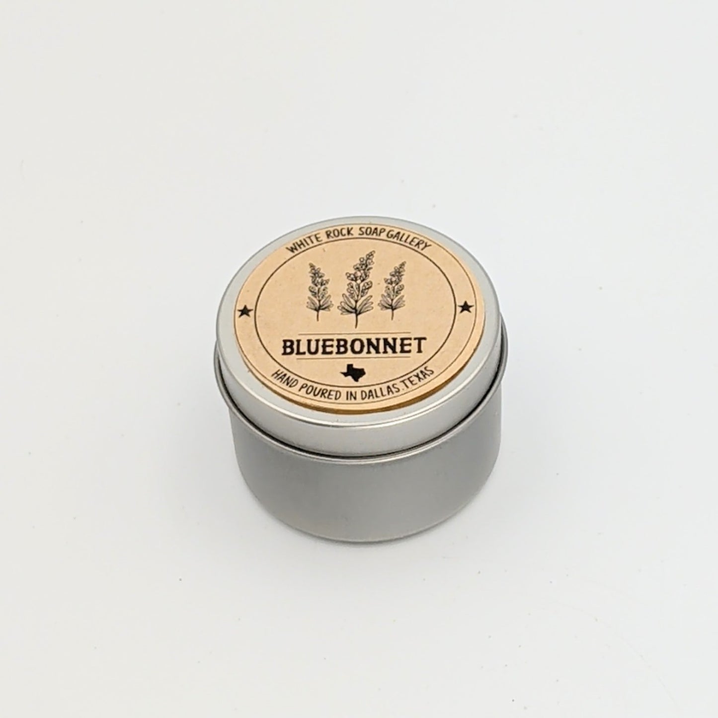 Silver metal candle tin labeled “Bluebonnet,” featuring a minimalist wildflower design and noting hand-poured craftsmanship in Dallas, Texas, displayed on a white background.