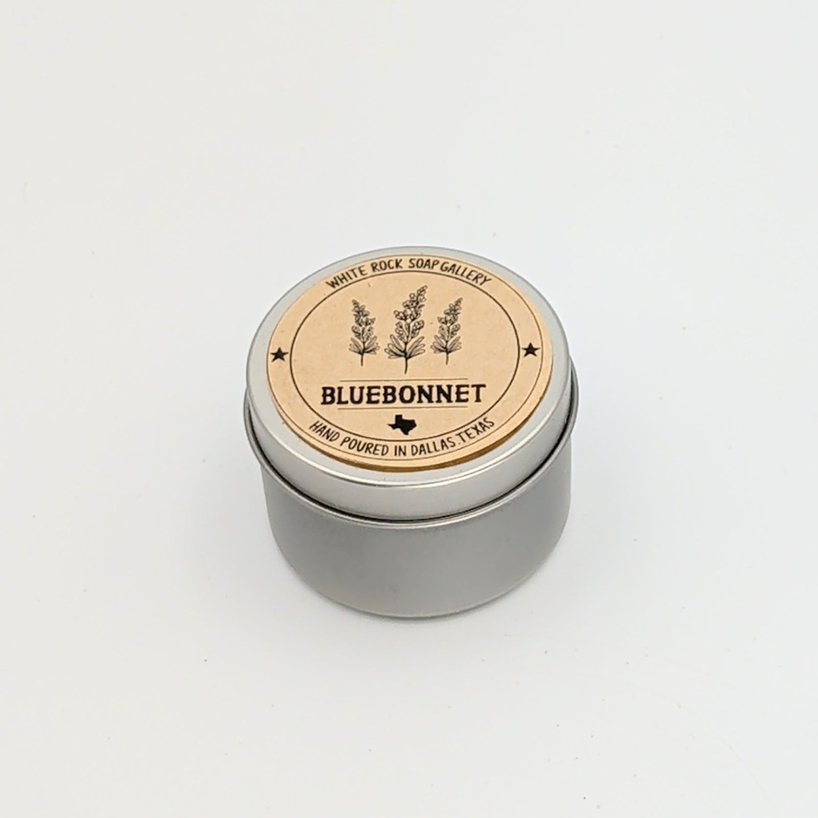 Silver metal candle tin labeled “Bluebonnet,” featuring a minimalist wildflower design and noting hand-poured craftsmanship in Dallas, Texas, displayed on a white background.