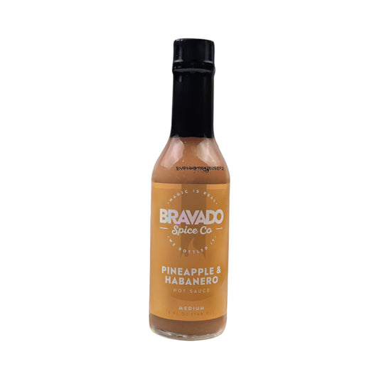 Bottle of Bravado Spice Co. Pineapple & Habanero hot sauce labeled “Medium,” featuring a golden-orange sauce in a clear glass bottle with a black cap, photographed on a white background