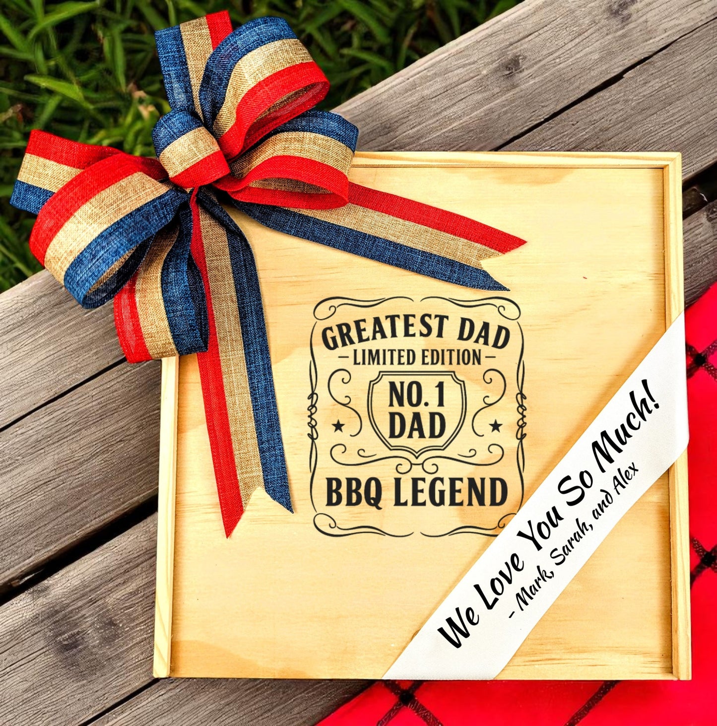 A wooden gift box with a custom lid engraving that reads 'Greatest Dad No. 1 BBQ Legend', with a red, blue, and beige ribbon on top, placed on a wooden surface with a plaid patterned fabric beneath it.