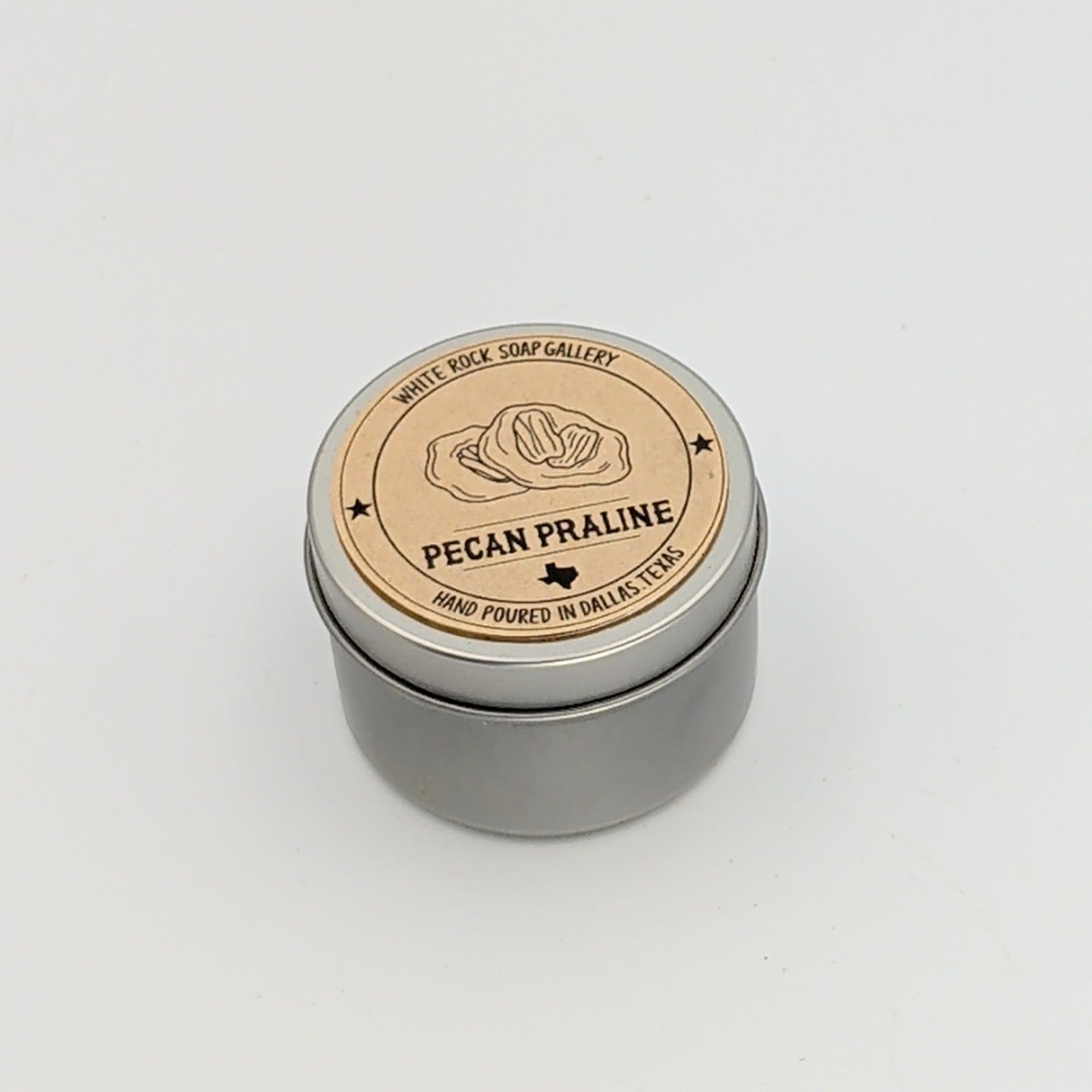 Silver metal candle tin labeled “Pecan Praline” by White Rock Soap Gallery, featuring a pecan illustration and noting hand poured in Dallas, Texas, photographed on a white background.