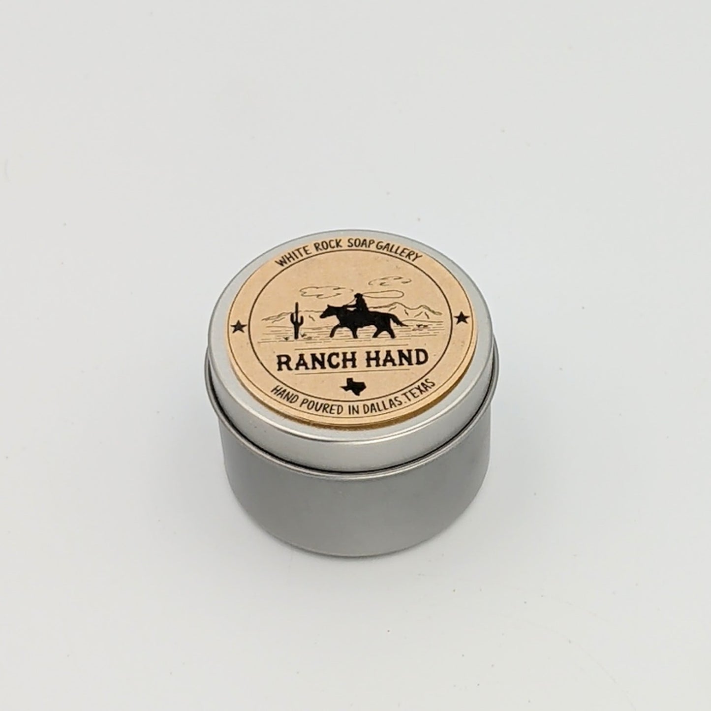 Silver metal candle tin labeled “Ranch Hand” with a silhouette of a cowboy on horseback and desert scenery, hand poured in Dallas, Texas, on a white background.