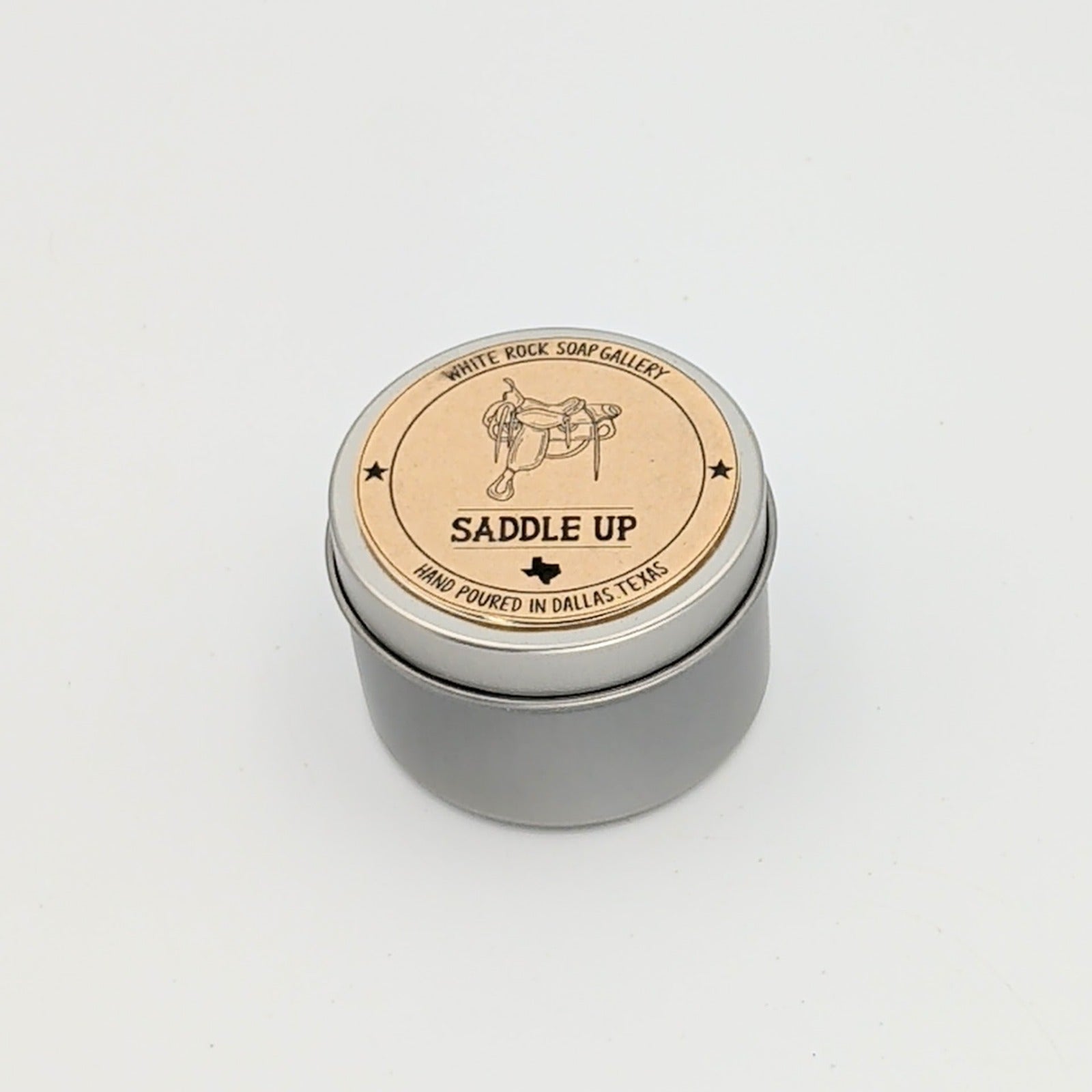 Silver metal candle tin labeled “Saddle Up” with a western saddle illustration, hand-poured in Dallas, Texas, photographed on a clean white background.